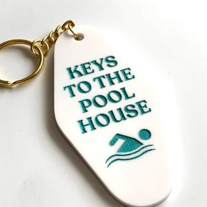 Op de afbeelding: Witte ovale sleutelhanger met goudkleurige ring en ketting. De sleutelhanger heeft de tekst "KEYS TO THE POOL HOUSE" in turquoise, met een zwemmer-icoon. Leuk accessoire.