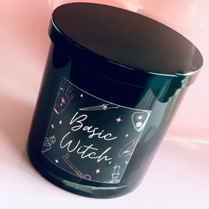 May include: Black candle jar with a pink and white label that says "Basic Witch." The label has illustrations of a hand with an eye, a knife, a crystal, a key, and a candle.