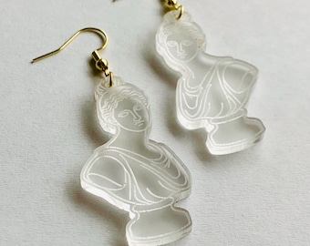 May include: A pair of clear acrylic earrings featuring a bust of a woman's face. The earrings have gold hooks.