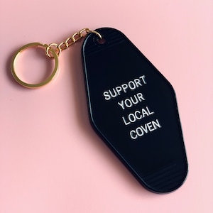 May include: Black motel key chain with the text "Support Your Local Coven" in white lettering.