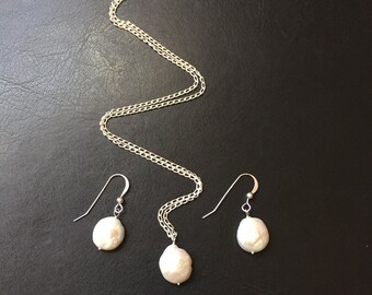May include: A set of pearl jewelry featuring a necklace and earrings. The necklace has a silver chain and a large, white pearl pendant. The earrings are silver with a white pearl dangling from each.