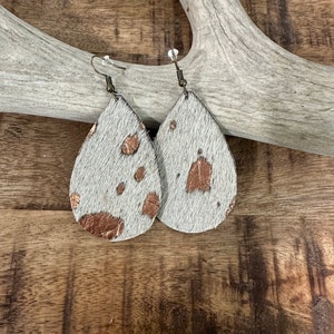 May include: A pair of teardrop-shaped earrings made of white cowhide with gold accents. The earrings have a simple hook closure.
