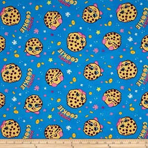 May include: A blue fabric with a repeating pattern of brown and white cookies with faces. The cookies have pink bows and are surrounded by gold coins, stars, and flowers. The text "Kooky Cookie" is repeated throughout the fabric.