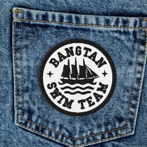 Pode incluir: Calças de ganga azul claro com um patch no bolso de trás. O patch é redondo, preto e branco, e apresenta um design de veleiro com as palavras "BANGTAN SWIM TEAM".