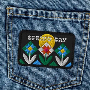 Puede incluir: Vaqueros de mezclilla azul claro con un parche decorativo en el bolsillo trasero. El parche presenta un diseño floral con las palabras "SPRING DAY" en letras blancas. Los vaqueros tienen un diseño clásico de cinco bolsillos.