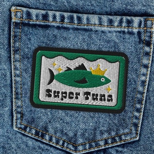 May include: Blue denim jeans with a green and white patch on the back pocket. The patch features a cartoon tuna wearing a crown and the text "Super Tuna".