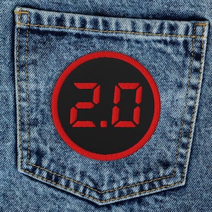 May include: Light blue denim jeans with a back pocket featuring a black patch with the red text "2.0" inside a red circle. The jeans have classic five-pocket styling and a button and zipper closure.