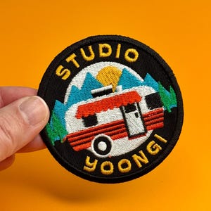 May include: Round embroidered patch with a black border featuring a red and white camper with the text "STUDIO YOONGI". The design includes blue mountains, green trees, and a yellow sun.