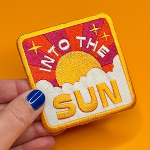 Puede incluir: Parche bordado con las palabras "INTO THE SUN" en blanco, amarillo y naranja. El parche presenta un diseño de sol con un fondo rosa y naranja y nubes blancas. El parche se sostiene sobre un fondo naranja.