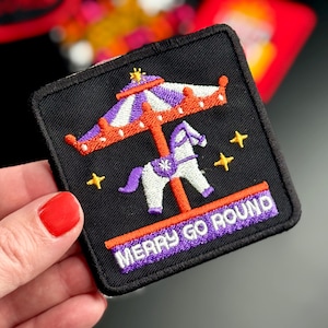 May include: A square black embroidered patch featuring a carousel horse with a purple mane and tail, a purple and white striped canopy, and orange accents. The text MERRY GO ROUND is embroidered in white on a purple background at the bottom.