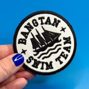 May include: Round embroidered patch with the words "BANGTAN SWIM TEAM" in black lettering on a white background. A black ship graphic is in the center. The patch has a black border and is on a blue background.
