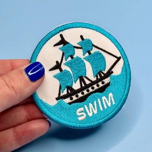 May include: Embroidered patch featuring a black and white ship with turquoise sails and water. The word "SWIM" is embroidered in white on the bottom. The patch is round with a turquoise border.