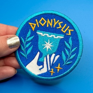 Könnte beinhalten: Ein runder, gestickter Aufnäher mit dem Wort "DIONYSUS" in Gelb. Er zeigt einen blauen Kelch, der von einer weißen Hand gehalten wird, mit einem Stern und goldenen Akzenten. Der Aufnäher hat einen blauen Rand und türkisfarbene Blätter.