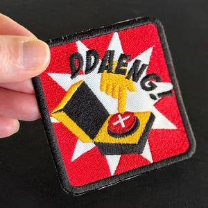 May include: Embroidered patch with the word "DDAENG!" in white, featuring a yellow hand pressing a red button. The patch has a red background with white starbursts and a black border.