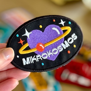 May include: An oval-shaped embroidered patch with a black background featuring a purple heart encircled by a yellow ring. The design includes small colourful stars and dots, with the white text MIKROKOSMOS printed across the lower section.