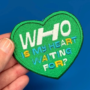 Pode incluir: Etiqueta verde em forma de coração com texto bordado branco que diz "WHO IS MY HEART WAITING FOR?" O texto está em várias cores, incluindo amarelo, azul e branco.