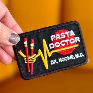 May include: Black rectangular patch with the words "PASTA DOCTOR" in white letters on a red circle. The patch features a yellow heartbeat line, a red and yellow pasta design, and the text "DR. KOOKIE, M.D." in white.