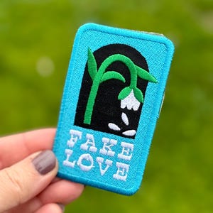 May include: A rectangular patch with a teal border and a black arched design featuring a green stem and white flower. The text "FAKE LOVE" is embroidered in white. The patch is held against a blurred green background.