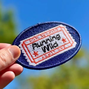 Könnte beinhalten: Ein blauer und weißer gestickter Aufnäher mit dem Text "Running Wild" in einem roten Sternenausbruch. Der Aufnäher ist ticketförmig und hat einen weißen Rand.