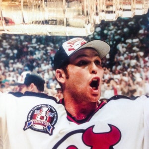 May include: A hockey player in a white jersey with a red and black logo, celebrating with the Stanley Cup. The jersey has a patch that says "Western Conference Eastern Stanley Cup 1995". The player is wearing a black and white hat.