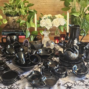 May include: A collection of black glass tableware including a pitcher, swan-shaped dish, cups, bowls, and plates arranged on a white tablecloth with star patterns. The display features candles, crystal accents, and potted plants in the background.