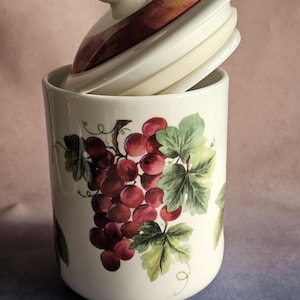May include: A cylindrical ceramic canister with a cream-colored base featuring a painted illustration of clusters of red grapes and green leaves. The canister includes a matching lid with a knob handle and a dark red band around the rim.