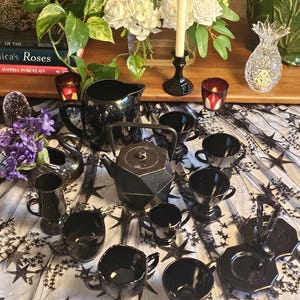 May include: A black tea set featuring a geometric teapot, a pitcher, and multiple cups with handles. The set includes a matching tray and is arranged on a white fabric surface patterned with black stars, alongside a swan-shaped vessel.
