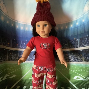 Puede incluir: Una mu&ntilde;eca vestida con una camiseta y pantalones rojos con el logotipo de los 49ers de San Francisco. La mu&ntilde;eca tambi&eacute;n lleva un gorro de punto granate con un pomp&oacute;n amarillo y zapatillas deportivas blancas y negras. El fondo es un estadio.