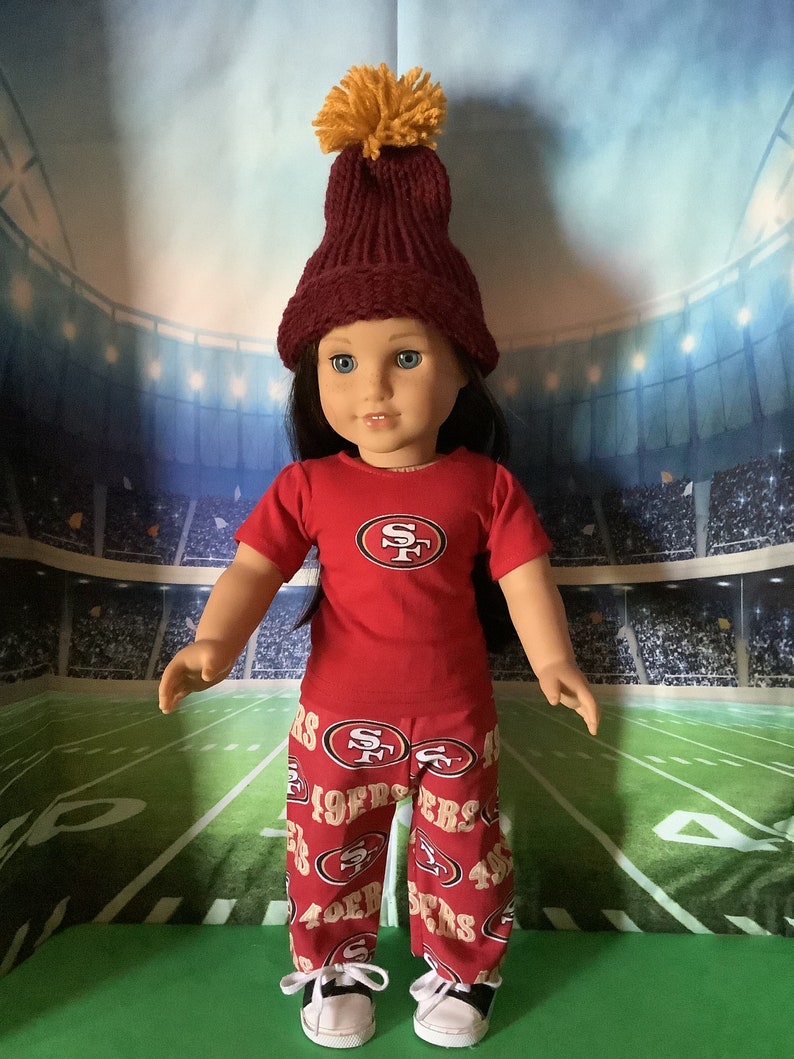 Puede incluir: Una mu&ntilde;eca vestida con una camiseta y pantalones rojos con el logotipo de los 49ers de San Francisco. La mu&ntilde;eca tambi&eacute;n lleva un gorro de punto granate con un pomp&oacute;n amarillo y zapatillas deportivas blancas y negras. El fondo es un estadio.