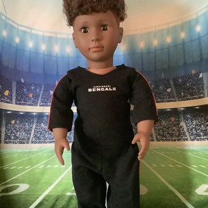 May include: A black and red football jersey with the text "Cincinnati Bengals" on the front. The jersey is being worn by a doll with brown curly hair.