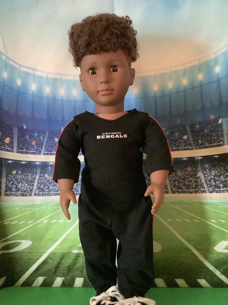 May include: A black and red football jersey with the text "Cincinnati Bengals" on the front. The jersey is being worn by a doll with brown curly hair.