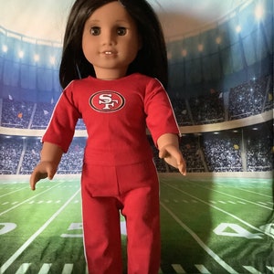 Puede incluir: Mu&ntilde;eca vestida con un atuendo deportivo rojo con rayas blancas y un logotipo del equipo en el pecho. La mu&ntilde;eca tiene cabello oscuro y usa zapatos rojos. El fondo es un campo de f&uacute;tbol.