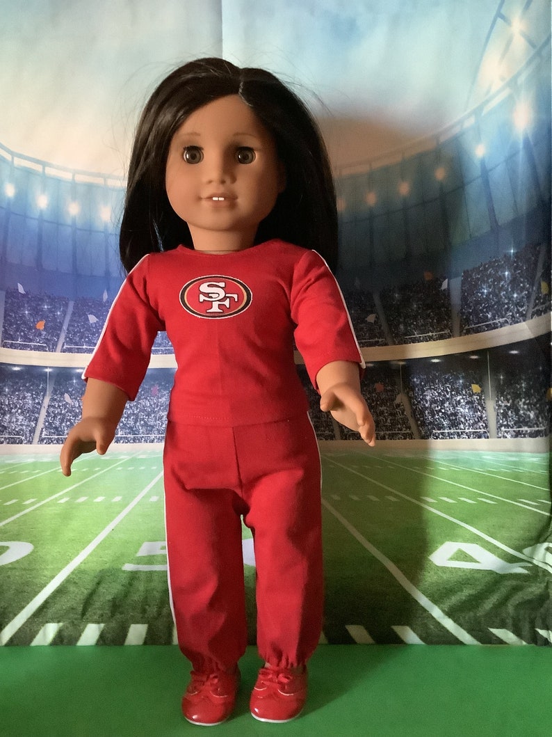 Puede incluir: Mu&ntilde;eca vestida con un atuendo deportivo rojo con rayas blancas y un logotipo del equipo en el pecho. La mu&ntilde;eca tiene cabello oscuro y usa zapatos rojos. El fondo es un campo de f&uacute;tbol.