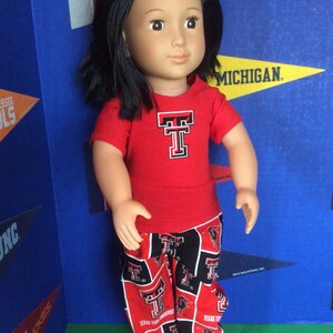 May include: A doll wearing a red t-shirt with a black and red patterned pants. The t-shirt has a large white "T" with a red outline. The pants have a repeated pattern of the "T" and a bull's head. The doll has long black hair and brown eyes.