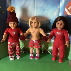 Puede incluir: Tres mu&ntilde;ecas vestidas con atuendos tem&aacute;ticos de los 49ers de San Francisco. Una mu&ntilde;eca lleva una camisa roja, pantalones y un gorro de punto con un pomp&oacute;n amarillo. Otra mu&ntilde;eca viste una camisa color beige y una falda roja. La tercera mu&ntilde;eca lleva un mono rojo.