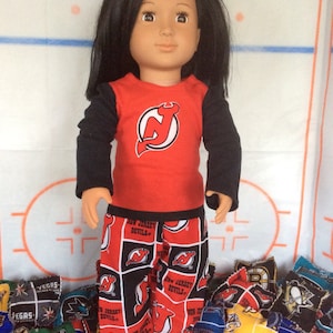 May include: A doll wearing a red long-sleeved shirt with a black New Jersey Devils logo and black and red pajama pants with the New Jersey Devils logo. The doll has long black hair and brown eyes. The doll is standing on a blue surface with a variety of colorful patches.