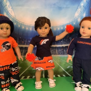 May include: Three dolls dressed in Denver Broncos themed outfits. The dolls are wearing orange and blue shirts and trousers with the Broncos logo. One doll is holding a red pom-pom.