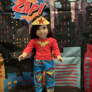 Puede incluir: Una mu&ntilde;eca vestida con un disfraz de Wonder Woman, que incluye una camiseta roja de manga larga con el logotipo de Wonder Woman, pantalones azules con logotipos de Wonder Woman y una diadema dorada con forma de estrella. El fondo presenta un paisaje urbano y la palabra "ZAP!"