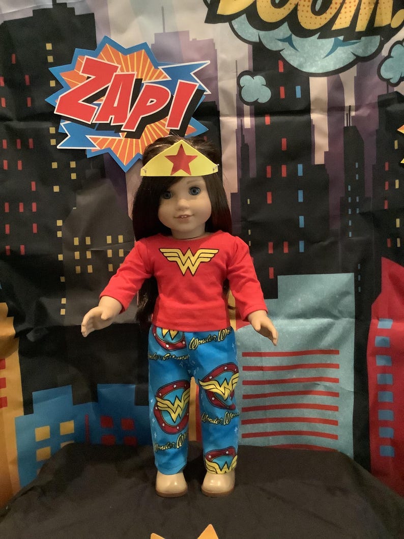 Puede incluir: Una mu&ntilde;eca vestida con un disfraz de Wonder Woman, que incluye una camiseta roja de manga larga con el logotipo de Wonder Woman, pantalones azules con logotipos de Wonder Woman y una diadema dorada con forma de estrella. El fondo presenta un paisaje urbano y la palabra "ZAP!"