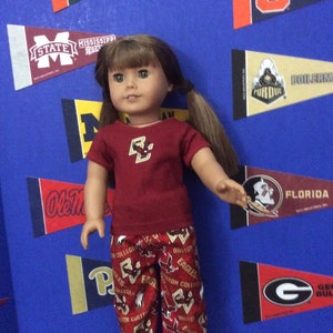 May include: A doll wearing a red shirt with a Boston College logo and red and gold patterned pants with the Boston College logo. The doll is standing in front of a wall with pennants from different universities.