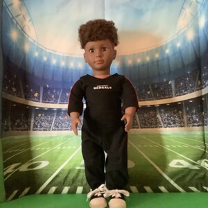 May include: A black and red doll wearing a black long-sleeved shirt with the words "Cincinnati Bengals" on the front. The doll is wearing black pants and white sneakers. The doll is standing in front of a green and white football field.