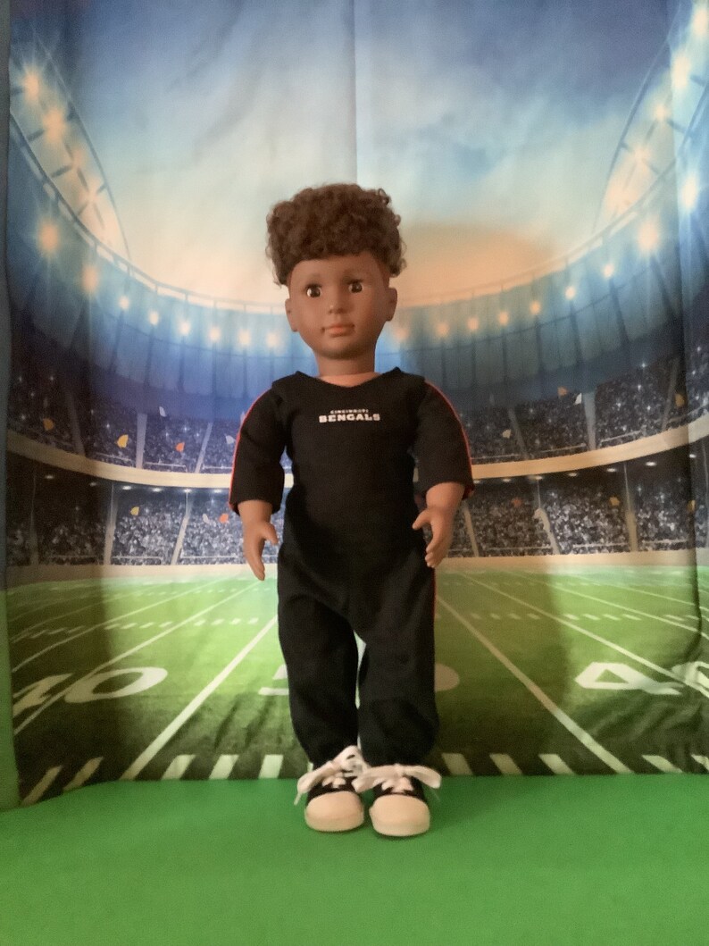 May include: A black and red doll wearing a black long-sleeved shirt with the words "Cincinnati Bengals" on the front. The doll is wearing black pants and white sneakers. The doll is standing in front of a green and white football field.