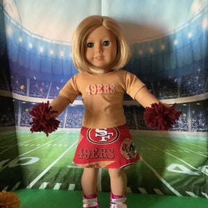 Puede incluir: Mu&ntilde;eca vestida con una camiseta dorada y una falda roja con el texto "49ERS". La mu&ntilde;eca tiene pelo rubio, zapatos rosas y sostiene pompones granates. El fondo es un campo de f&uacute;tbol americano.