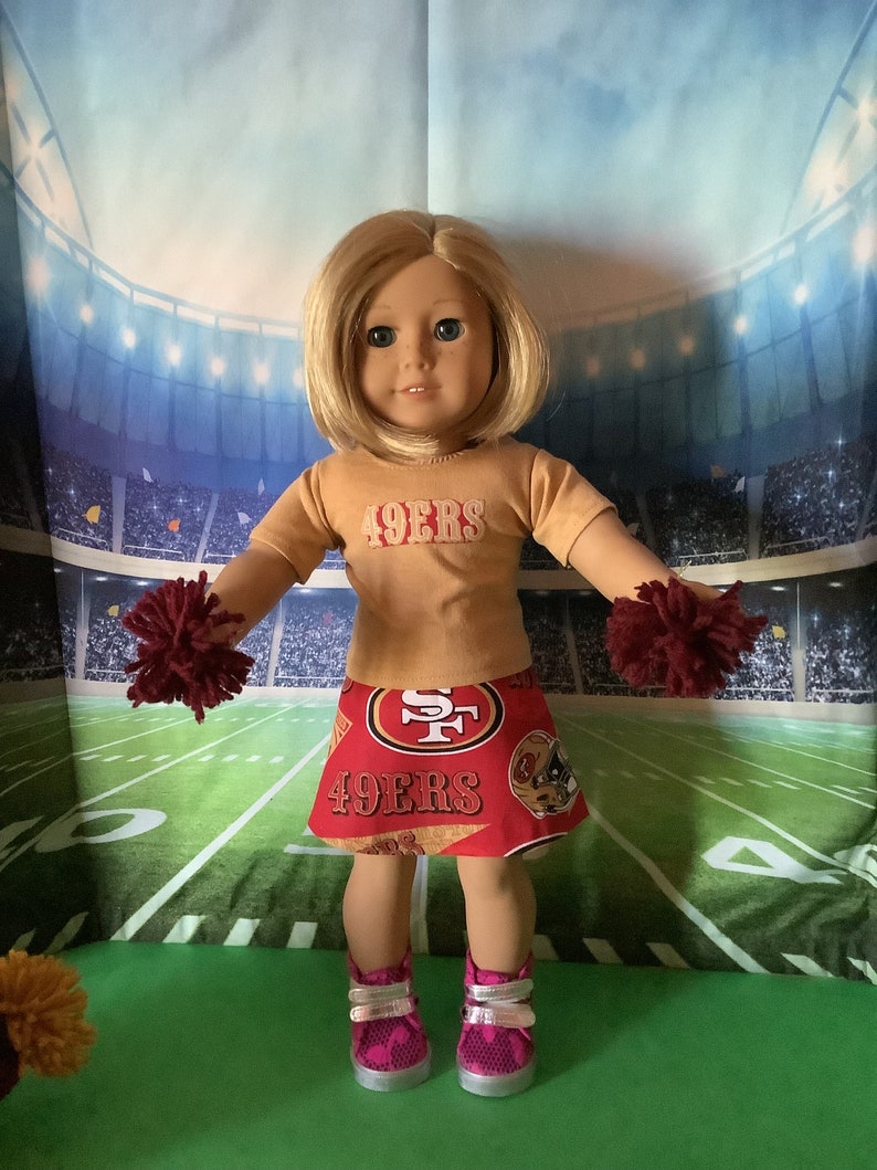 Puede incluir: Mu&ntilde;eca vestida con una camiseta dorada y una falda roja con el texto "49ERS". La mu&ntilde;eca tiene pelo rubio, zapatos rosas y sostiene pompones granates. El fondo es un campo de f&uacute;tbol americano.