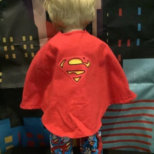 Puede incluir: Una capa roja de Superman con un logotipo amarillo y negro se usa sobre un conjunto de pijama de Superman azul y rojo. El conjunto de pijama presenta un gr&aacute;fico de Superman. El fondo tiene un horizonte de la ciudad.