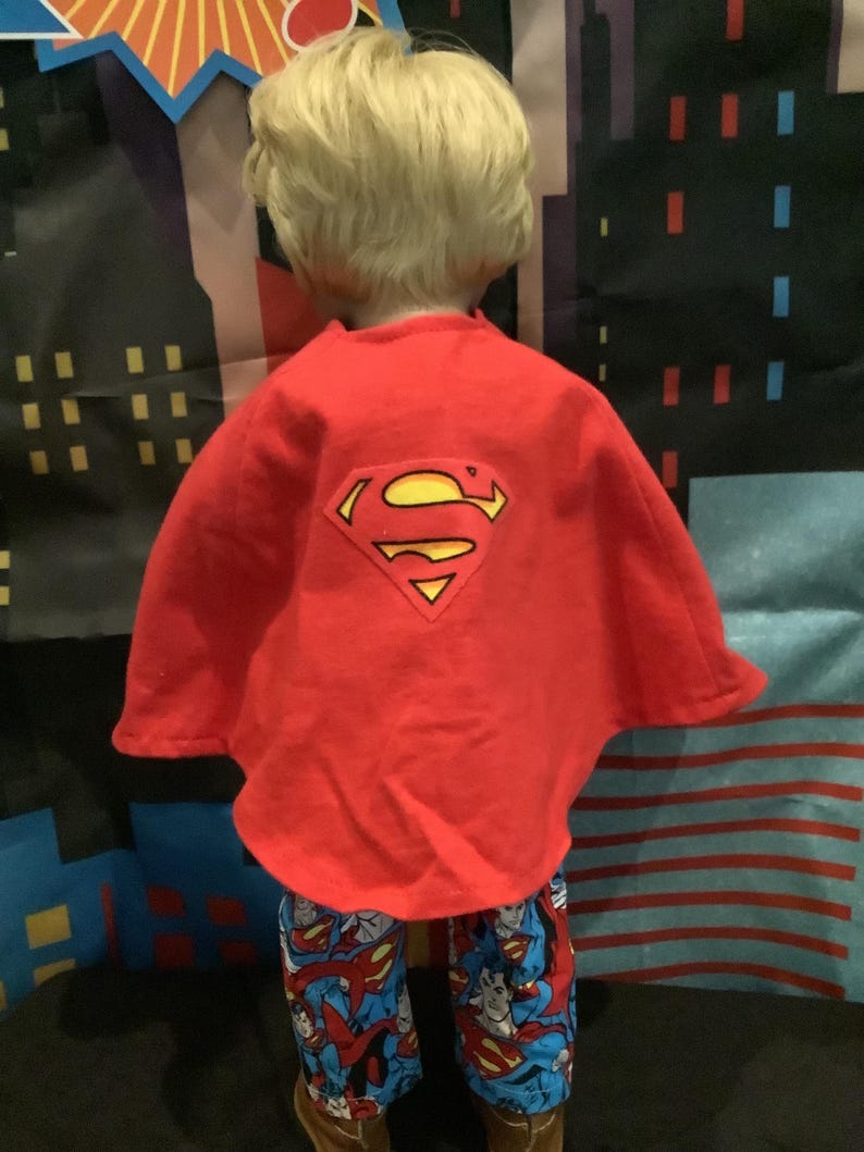 Puede incluir: Una capa roja de Superman con un logotipo amarillo y negro se usa sobre un conjunto de pijama de Superman azul y rojo. El conjunto de pijama presenta un gr&aacute;fico de Superman. El fondo tiene un horizonte de la ciudad.