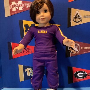 May include: A doll wearing a purple and gold LSU tracksuit with white sneakers. The doll has brown hair and blue eyes.