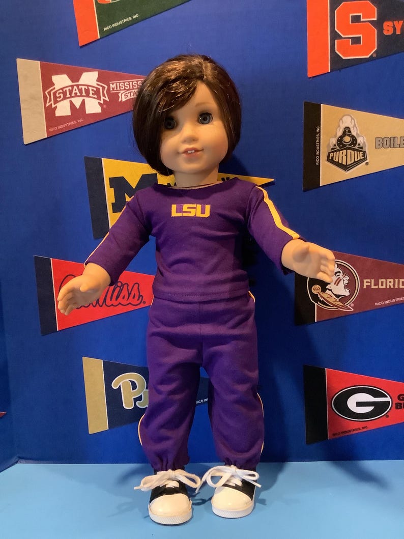 May include: A doll wearing a purple and gold LSU tracksuit with white sneakers. The doll has brown hair and blue eyes.