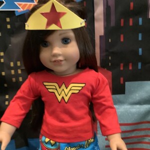 Puede incluir: Una mu&ntilde;eca vestida como Wonder Woman, con una camisa roja de manga larga con un emblema dorado y pantalones azules estampados. La mu&ntilde;eca tiene una corona amarilla con una estrella roja. El fondo presenta un paisaje urbano.