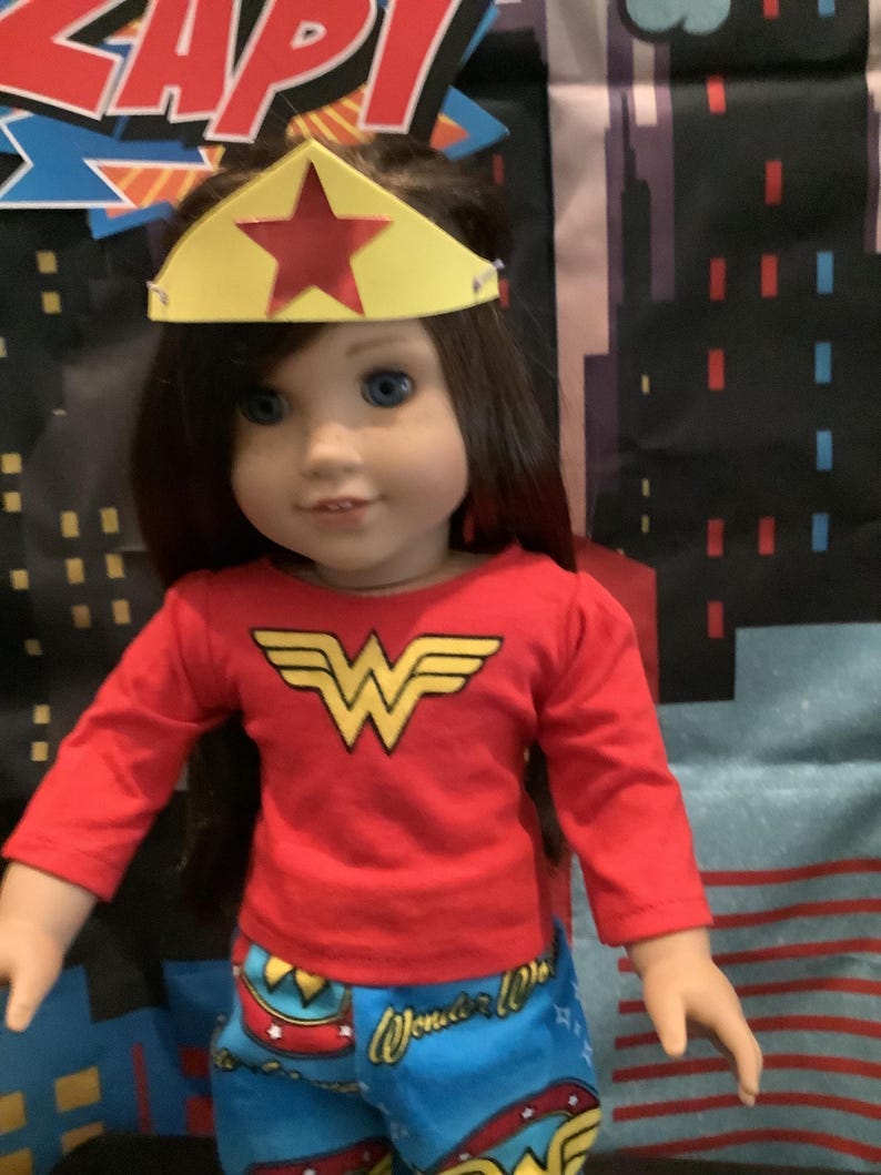 Puede incluir: Una mu&ntilde;eca vestida como Wonder Woman, con una camisa roja de manga larga con un emblema dorado y pantalones azules estampados. La mu&ntilde;eca tiene una corona amarilla con una estrella roja. El fondo presenta un paisaje urbano.
